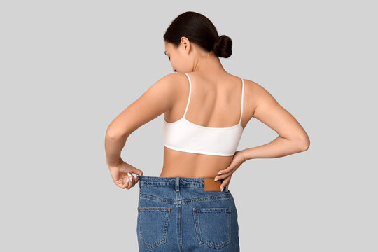 Young woman in loose jeans after weight loss on light background, back view