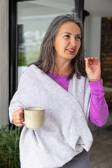 Middle-aged woman wearing shawl standing near sliding glass door holding ceramic mug and gesturing © wavebreak3