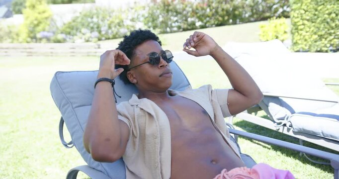 African-American man reclining on garden chaise in shirt feeling sun adjusting shades and settling