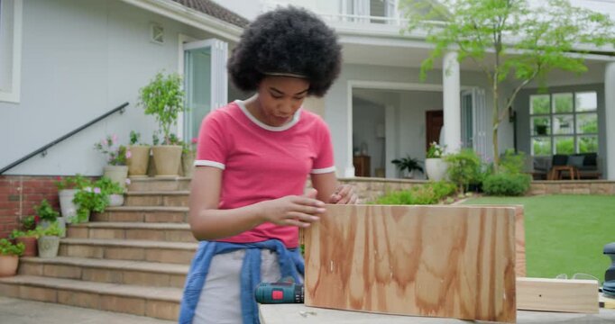 African American woman checking phone, using drill fastening plywood box on patio bench in pink tee