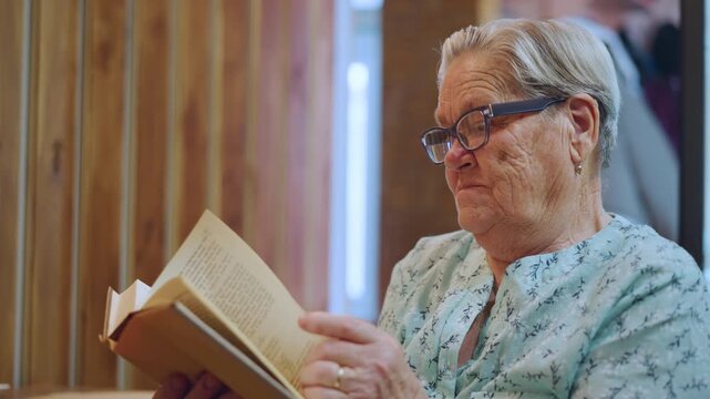 Elderly woman reading. Grandmother gazes quietly indoors. Senior lady relaxes by cozy wooden wall. Senior woman reading in peaceful setting illuminated softly by home interior lighting