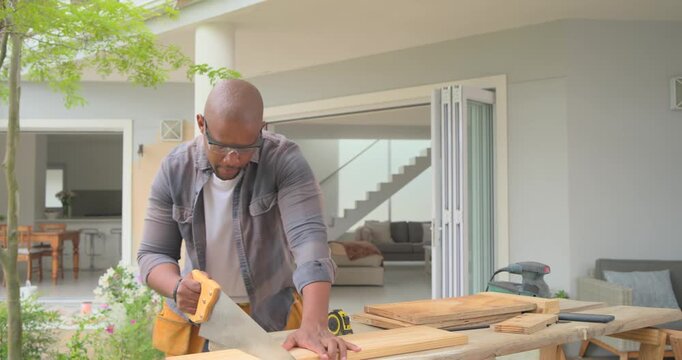 African American man wearing goggles placing planks, measuring, sizing boards with handsaw on patio