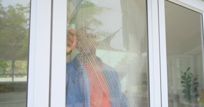 African man wearing denim shirt cleaning soapy sliding door at home, spraying and squeegeeing