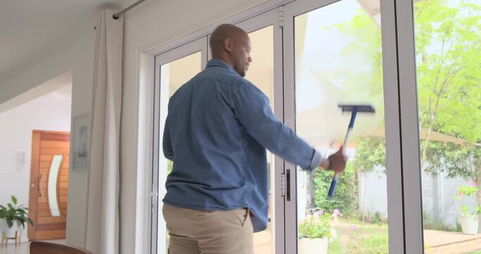 Entering frame, African man seeing streaks and cleaning glass door at home with squeegee in denim