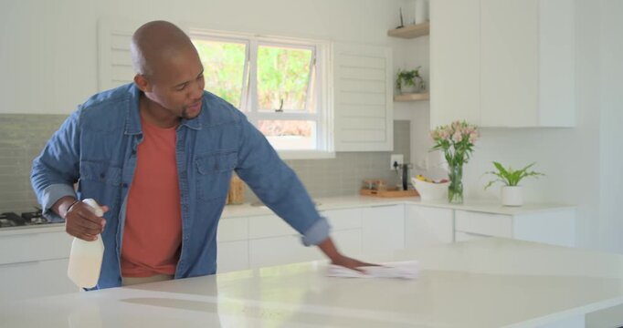 Middle-aged African man grabbing spray bottle spraying wiping island in kitchen with cloth to clean