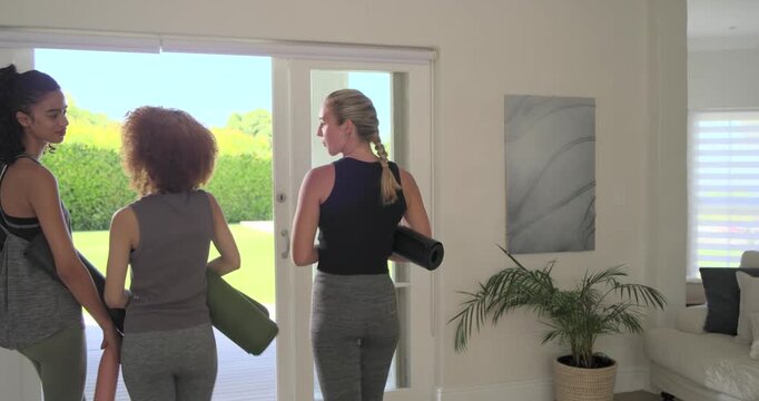 Diverse female friends walking to doorway in activewear holding mats, bottles, coordinating yoga