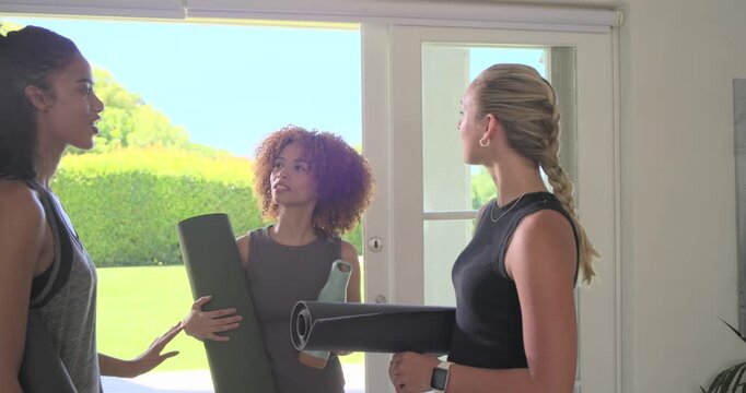 Diverse female friends holding rolled mats, bottle at glass door, left woman proposing yoga plan