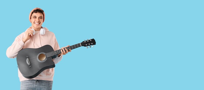 Happy young man with headphones and guitar on light blue background with space for text