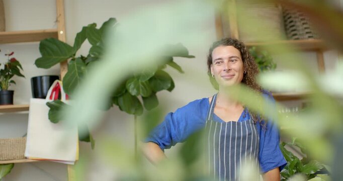 Non-binary seller in apron in plant shop greeting clients while camera pulling focus through leaves