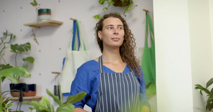 Non-binary attendant in plant shop wearing striped apron, shifting gaze as camera reframing plants