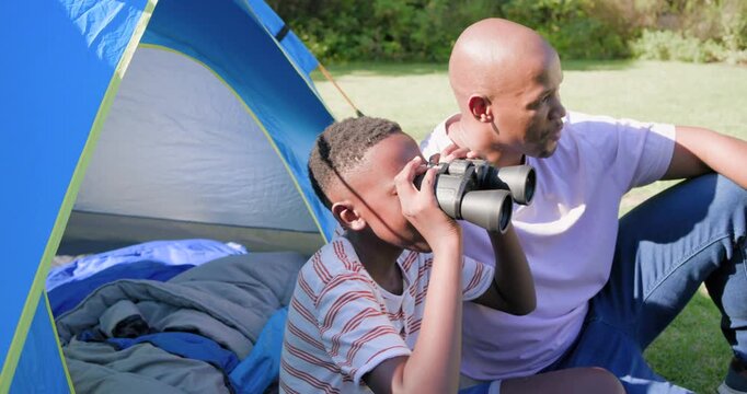 African American father and son sitting on grass by tent dad pointing guiding son using binoculars