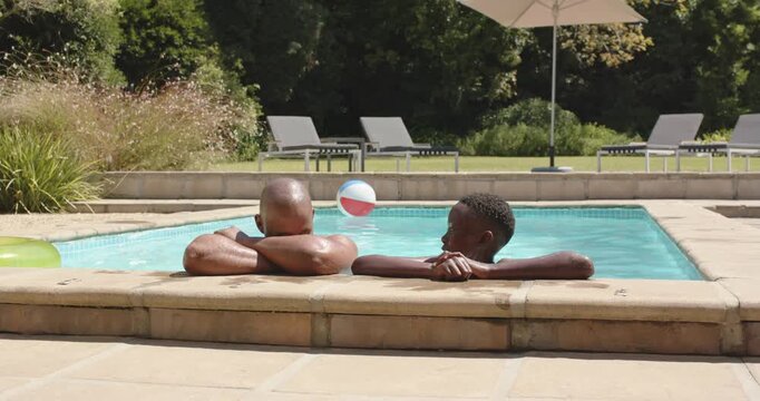 Adult male and youth male leaning on backyard pool edge, following airborne beach ball, playing