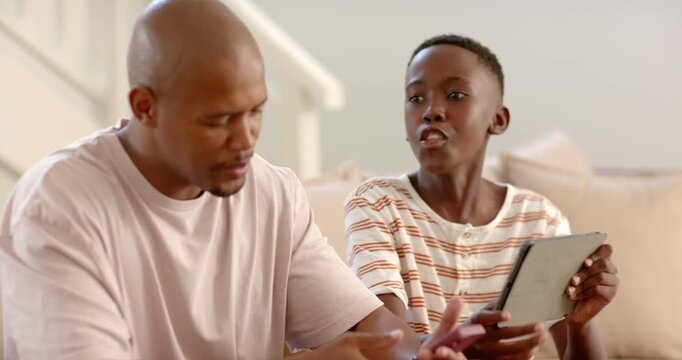 African father and son talking on couch as father reaching phone, son showing tablet, proving point
