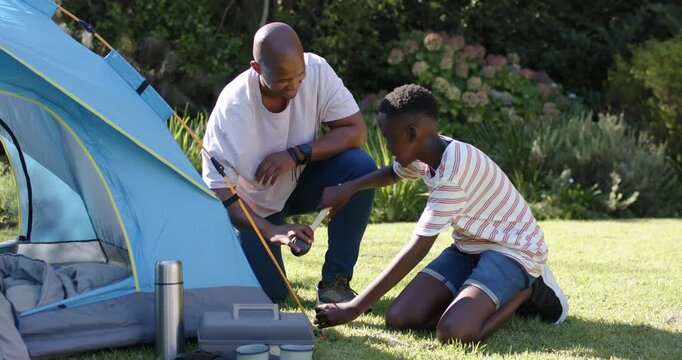 African American dad kneeling pointing teen son hammering stake with mallet securing tent on lawn