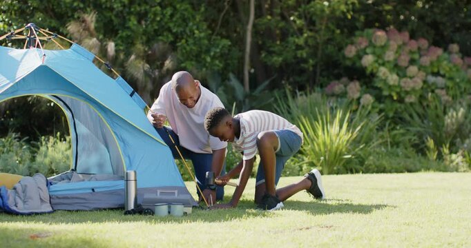 Father and son finding loose guy line, kneeling, hammering stake and securing blue tent in backyard