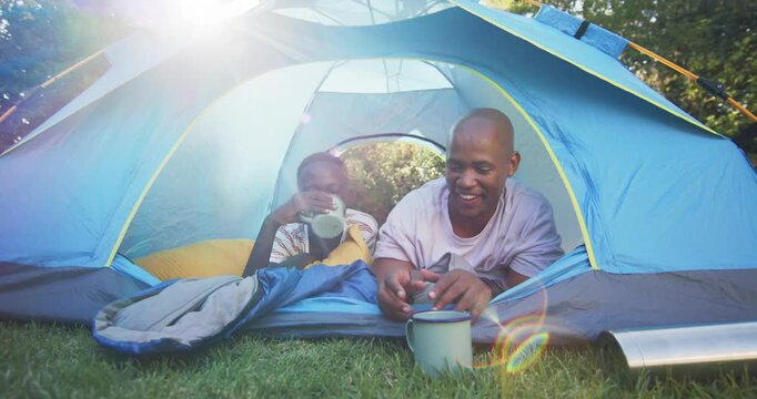 Sunlight waking African American father and son, peeking from blue tent, reaching mugs to drink