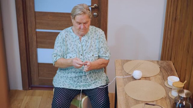 Grandma placing egg on wooden table, caucasian elderly woman concentrating on small task with steady reaching motion toward woven placemat, cup and kettle arranged nearby, warm interior light, focused