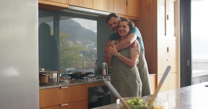 Cooking mature couple wearing aprons embracing in home kitchen, showing salad bowl and cooktop