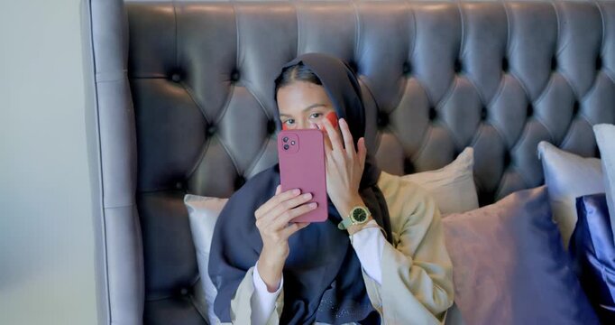 Woman in hijab holding pink smartphone using mirror aligning red patches for skincare on bed