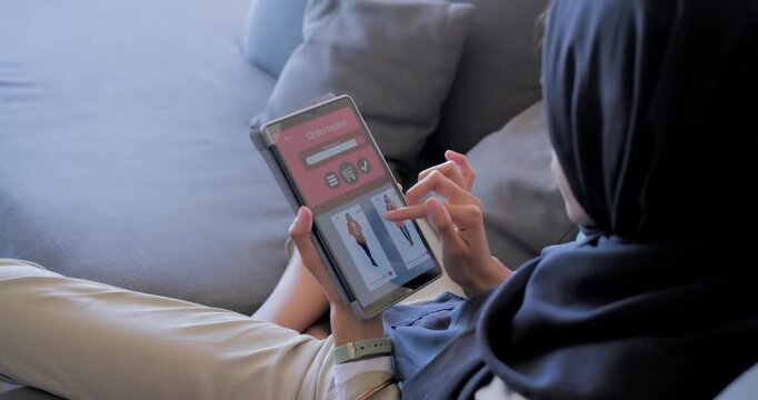 Woman lying on sofa in hijab, scrolling tablet showing thumbnails and viewing coat, copy space