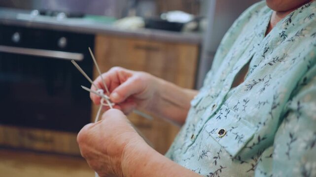 Closeup caucasian hands knitting with needles, kitchen background hints at cozy home routine, knuckles and fingers show practiced dexterity while wool loops form intricate stitch work with gentle