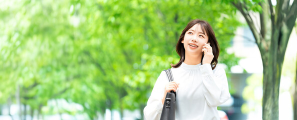 Fototapeta premium 遊歩道でスマートフォンで話す日本人女性・ビジネスウーマンのビジネスイメージ（LP・バナー向け）, Japanese Businesswoman Talking on Smartphone on Promenade for Landing Page Banner