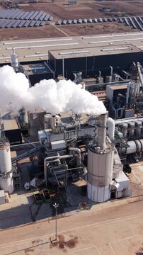 Vertical screen Aerial Industrial Site With Steam Plumes And Adjacent Solar Arrays, Contrast Between Heavy Industry And Renewable Energy, Storage Silos And Service Lanes, Bright Daylight