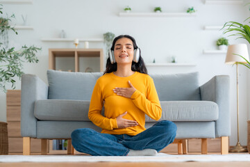 Obraz premium Happy young Asian woman practicing yoga and meditation at home sitting on couch in living room in lotus position and relaxing with closed eyes. Mindful meditation and wellbeing concept