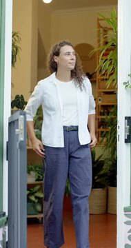 Vertical video: Walking nonbinary adult in white tee pants at plant shop door pausing to look right