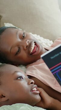 Vertical video: Opening striped book older sis reading to little sis to laugh on sofa in pink top