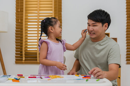 happy child girl playing and painting on her father's face with oil pastels