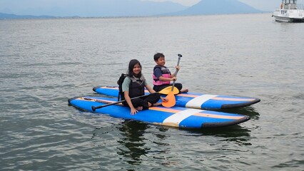 Obraz premium Exited East Asian kids are sitting on stand up paddle board at a beautiful lake under blue sky on with amazing hills and mountain in background. 
