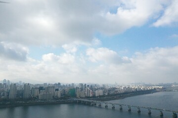Obraz premium Wide aerial shot of a city skyline beside a river under a soft cloudy sky.