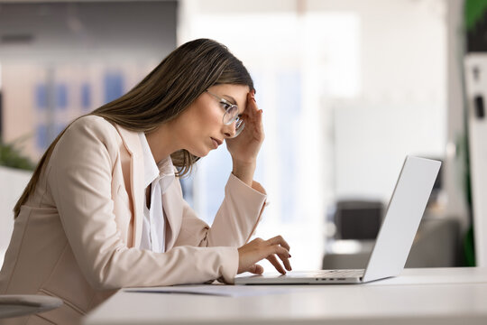 Concerned young businesswoman in glasses holding head while reading negative news, looking at laptop screen, feels exhausted, experiencing business failure, stress at workplace or business problems