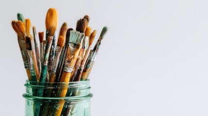 Fototapeta premium Close-up of paintbrushes in a clear, teal jar against a clean white background, showing their various sizes and stained bristles