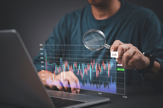 Male investor holding magnifying glass analyzing virtual stock market candlestick chart on laptop. Concept of financial data research, technical trading analysis, and investment audit strategy.