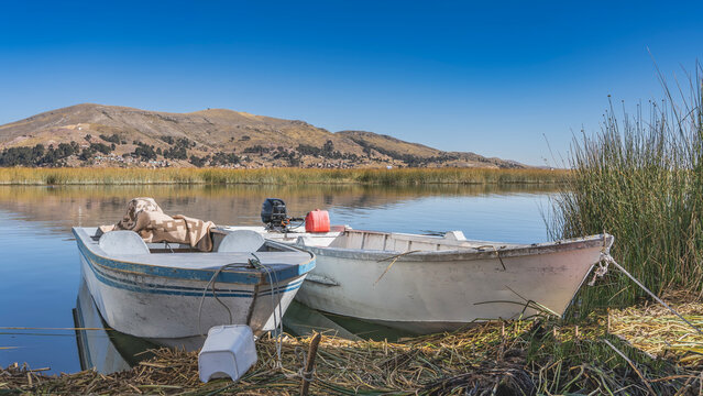 Two old motorboats are moored off the shore Uros Floating Island. Mountains against a blue sky. Thickets of totora reeds near the coast. Reflections on calm water. Peru. Lake Titicaca.