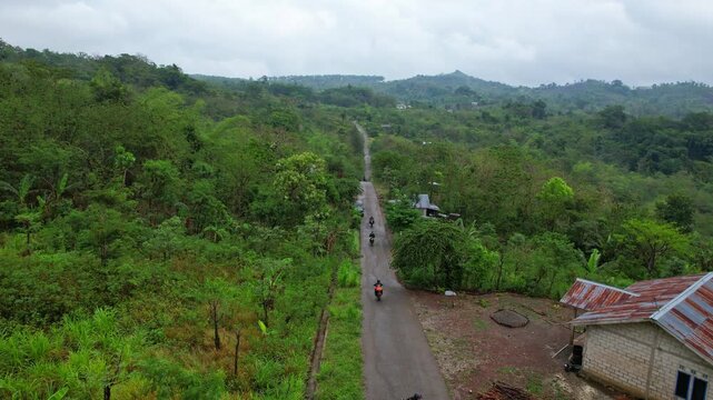 Aerial drone footage of motorbike rider group going thru a local long road, with wild forests and mountains behind, in Kupang regency, Timor island, Indonesia