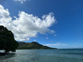 Obraz premium A serene tropical coastline with lush green mountains under a bright blue sky with fluffy white clouds.