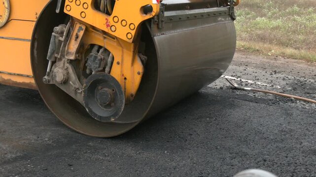 Roadworks, process of asphalting, blacktopping and paving new road layer. The metal cylinder of the yellow road vibrating roller compacts the fresh asphalt powerfully on the construction site
