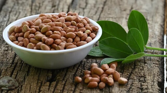 Raw peanuts in white bowl with green leaves on rustic wooden background