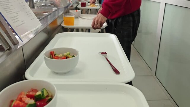 A person carrying a white tray with a fresh vegetable salad and a fork in a cafeteria or dining hall setting.