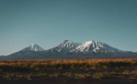 Panoramic landscape of three famous volcanoes on the Kamchatka Peninsula in Russia &mdash; Koryaksky, Avachinsky and Kozelsky. Snow-covered volcanic peaks rising above wild grassland under clear blue sky in
