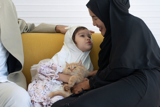 Happy Muslim family at home. Cute little girl in white hijab playfully pouts at her mother while holding a fluffy orange cat on a yellow sofa. Islamic lifestyle, pet care and family bonding.