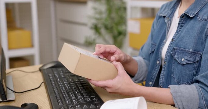 Asian woman small business owner rotating parcel box near computer desk while reading shipping label during e-commerce packaging process showing online seller customer order details before delivery