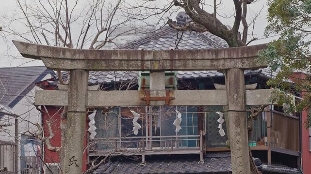 A static medium shot of a weathered stone torii gate in Japan, featuring paper streamers and a background of traditional tiled roofs and trees.