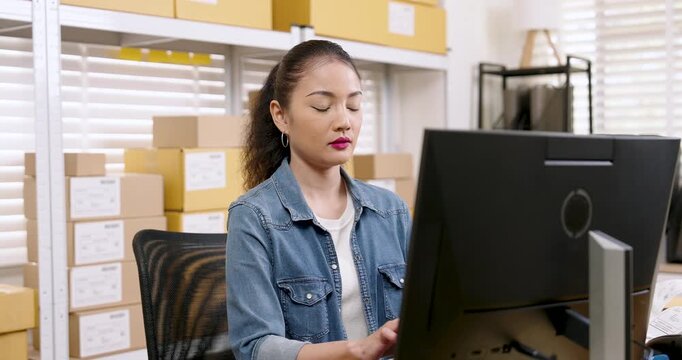 Asian woman small business owner working at desktop computer checking customer orders while online shopping parcel box stacks remain organized behind shelves showing e-commerce business workflow