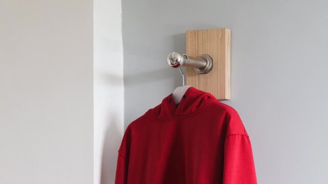 Hand hanging red hoodie on wall hanger inside home, minimalist interior clothing organization concept with metal hook and clean background.