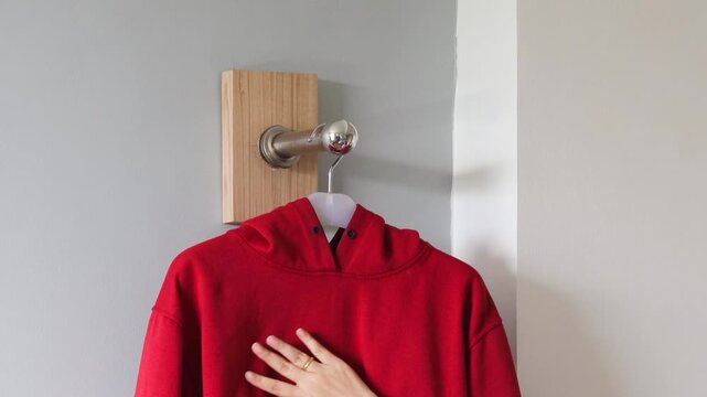 Hand hanging red hoodie on wall hanger inside home, minimalist interior clothing organization concept with metal hook and clean background.