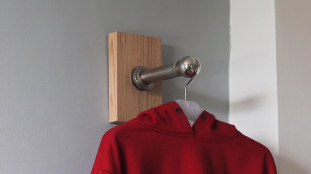 Hand hanging red hoodie on wall hanger inside home, minimalist interior clothing organization concept with metal hook and clean background.